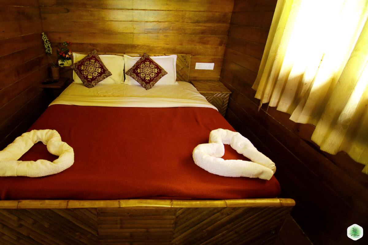 A wooden cabin-style room with a bed, two decorative pillows, a red bedspread, and two towels arranged on the bed.