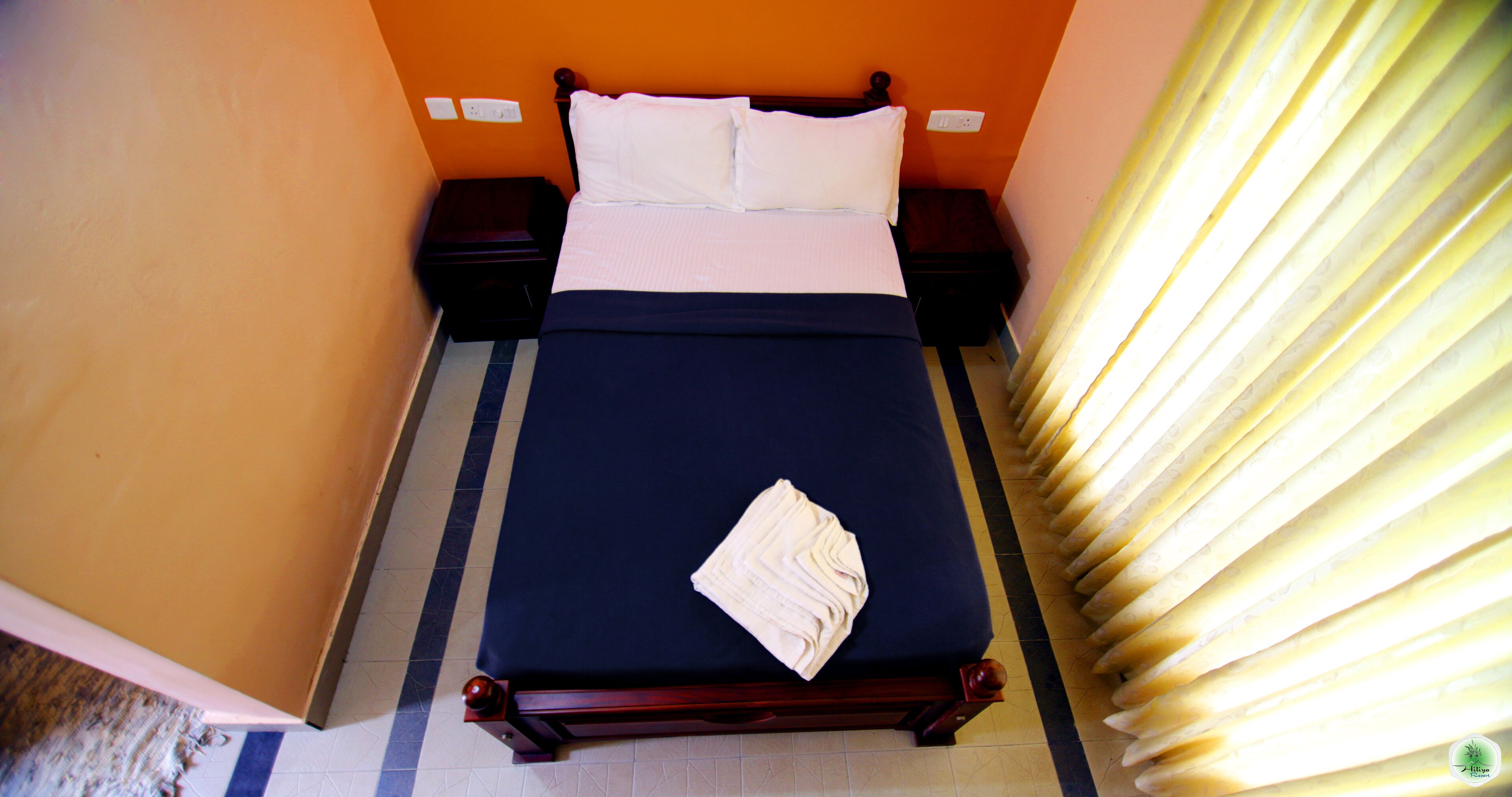 Un lit bien fait, recouvert d’une couvre-lit bleu foncé et agrémenté d’oreillers blancs, une serviette blanche pliée posée sur le lit, et des tables de chevet en bois à la finition sombre.