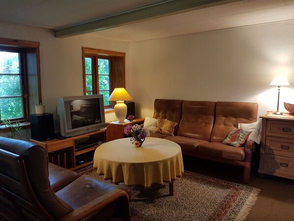 A living room with a brown leather sofa, a television on a wooden stand, a round coffee table with a white tablecloth, and a lamp on a side table.