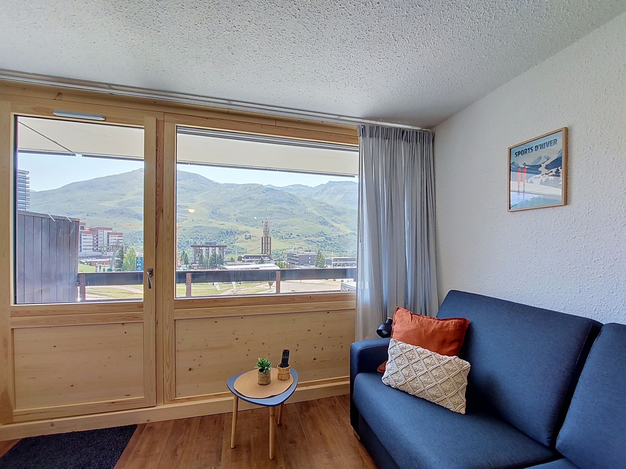 A room with a large window, a blue sofa, a small table with a plant, and a picture on the wall.