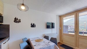 Un espace repas comprenant une table, des chaises, un téléviseur fixé au mur et une vue sur l’extérieur, visible à travers une porte vitrée.