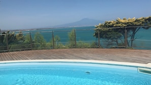 A swimming pool with clear blue water, surrounded by a wooden deck and glass railing, overlooking a scenic view of a mountain and a body of water. - New terrace sea view villa (Augusta)