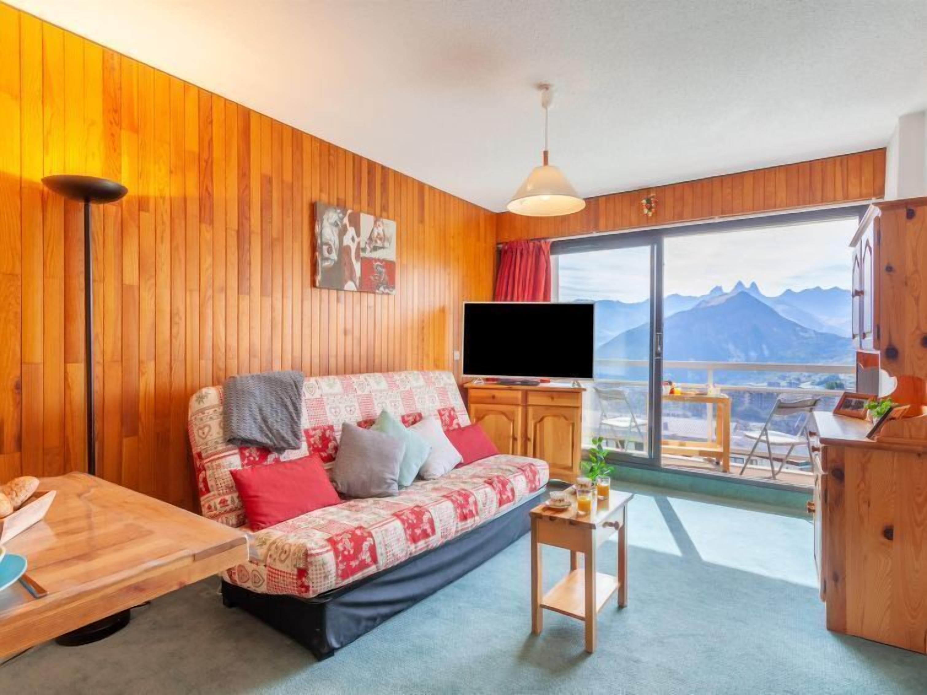 Quarto aconchegante com parede de madeira, sofá com manta estampada, televisão e vista para montanhas através de grandes janelas.