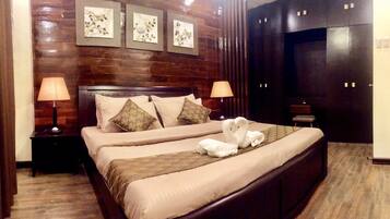 Habitación de hotel con cabecera de madera, dos lámparas de noche, cama con ropa de cama color beige y almohadas decorativas, una decoración de cisne blanco mullido, y un armario oscuro de dos puertas.