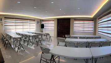 Un salón de conferencias con mesas y sillas blancas, ventanas amplias con persianas y un techo moderno con iluminación empotrada.