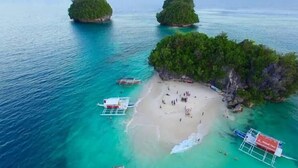 Une vue aérienne d’une plage aux eaux turquoise et limpides, bordée d’un rivage sablonneux, où l’on aperçoit plusieurs bateaux.