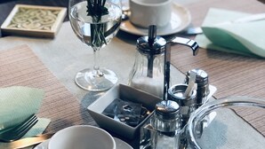 Una mesa de comedor ordenada con una taza blanca, un platito, una cuchara, un vaso de agua, salero y pimentero, un pequeño florero con flores blancas y una cafetera.