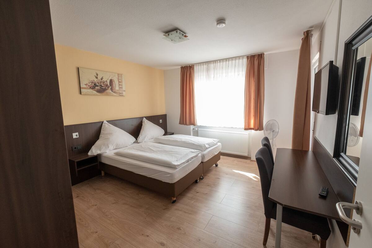 Habitación de hotel con cama, escritorio, silla, televisor y ventana con cortinas.