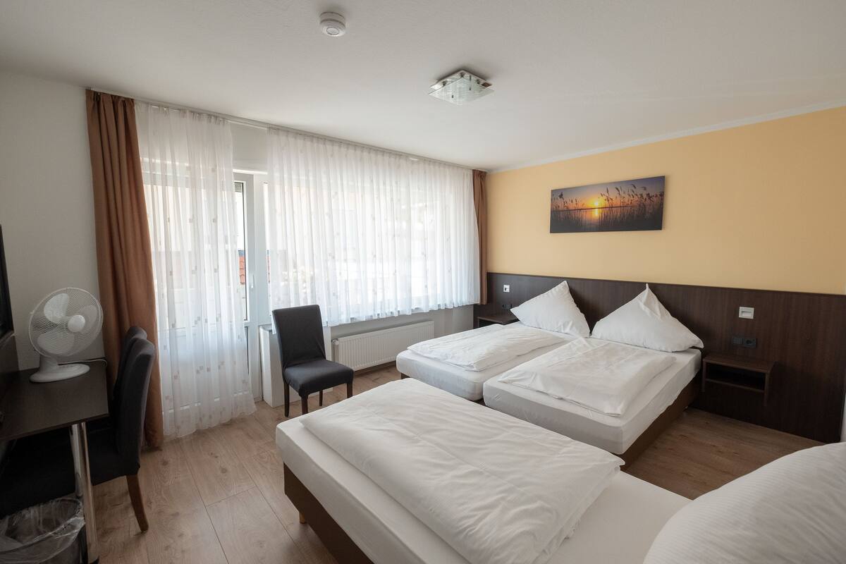Habitación de hotel con dos camas, un escritorio con un ventilador, una silla y una ventana con cortinas.