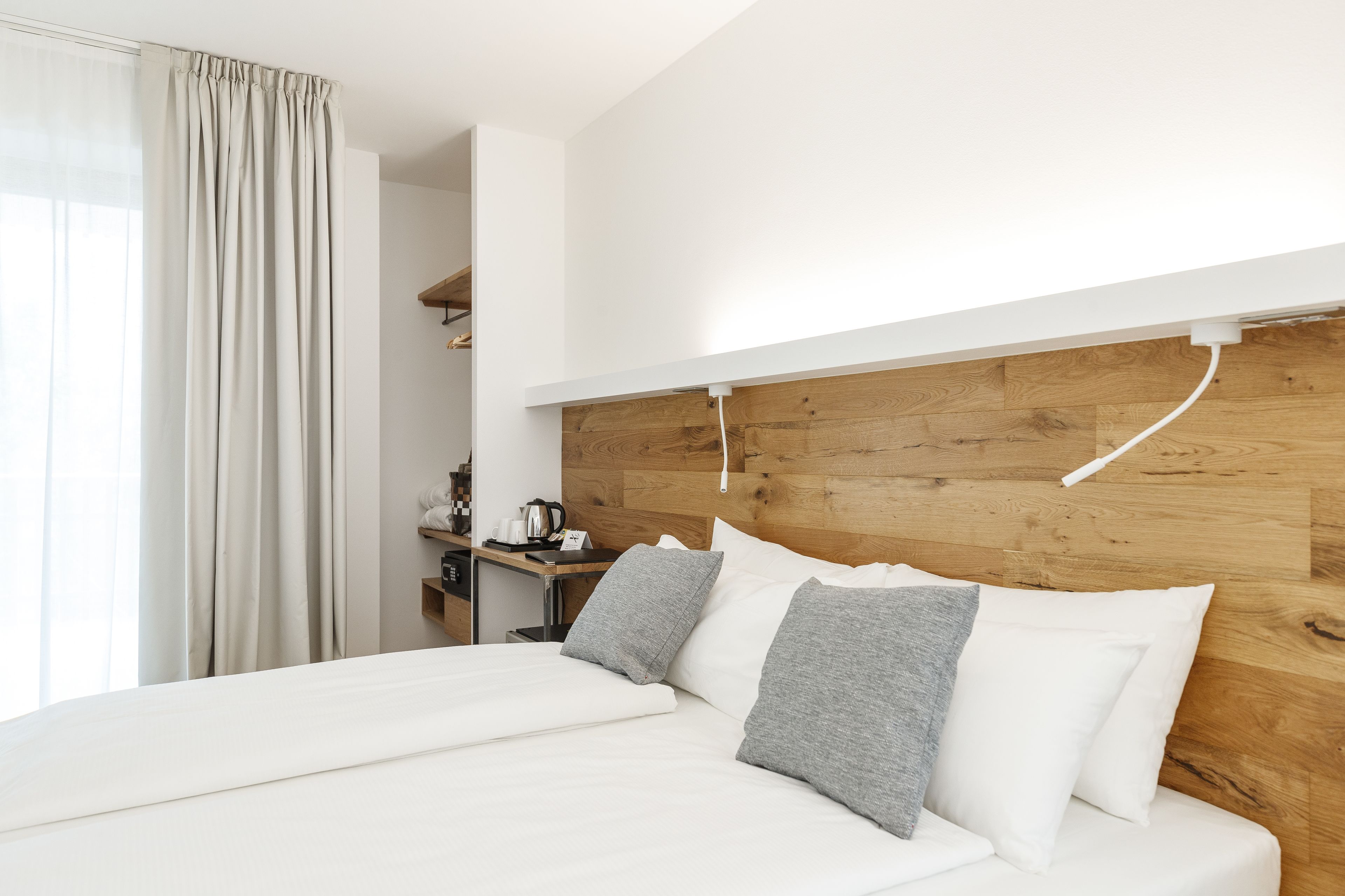 A modern hotel room with a wooden headboard, a neatly made bed with white and grey bedding, a small desk with a kettle, and a window with curtains.