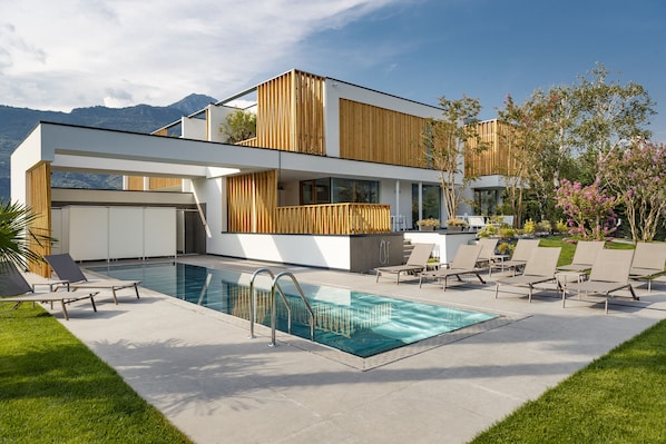 Una casa moderna de dos pisos con piscina, mobiliario exterior y montañas al fondo.