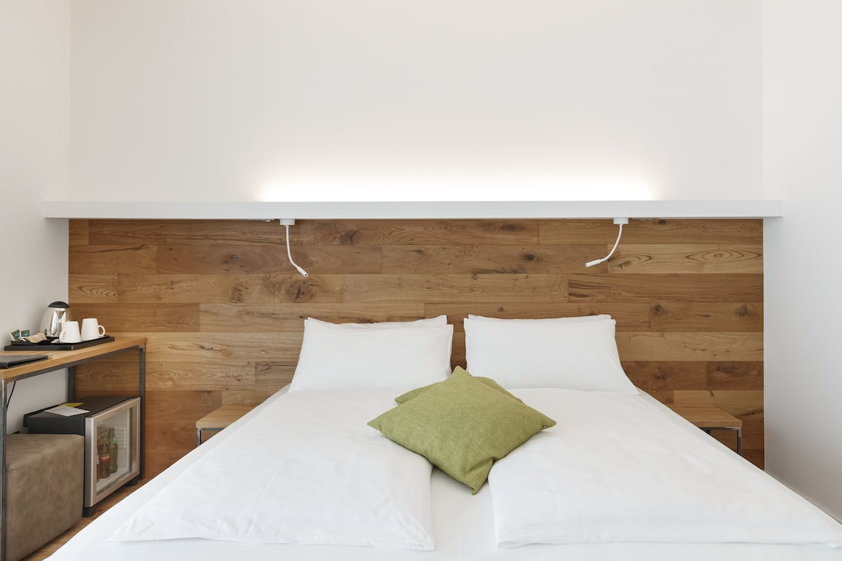 A neatly made bed with white linens and a green pillow, a wooden headboard, and a small bedside table with a kettle and cups.