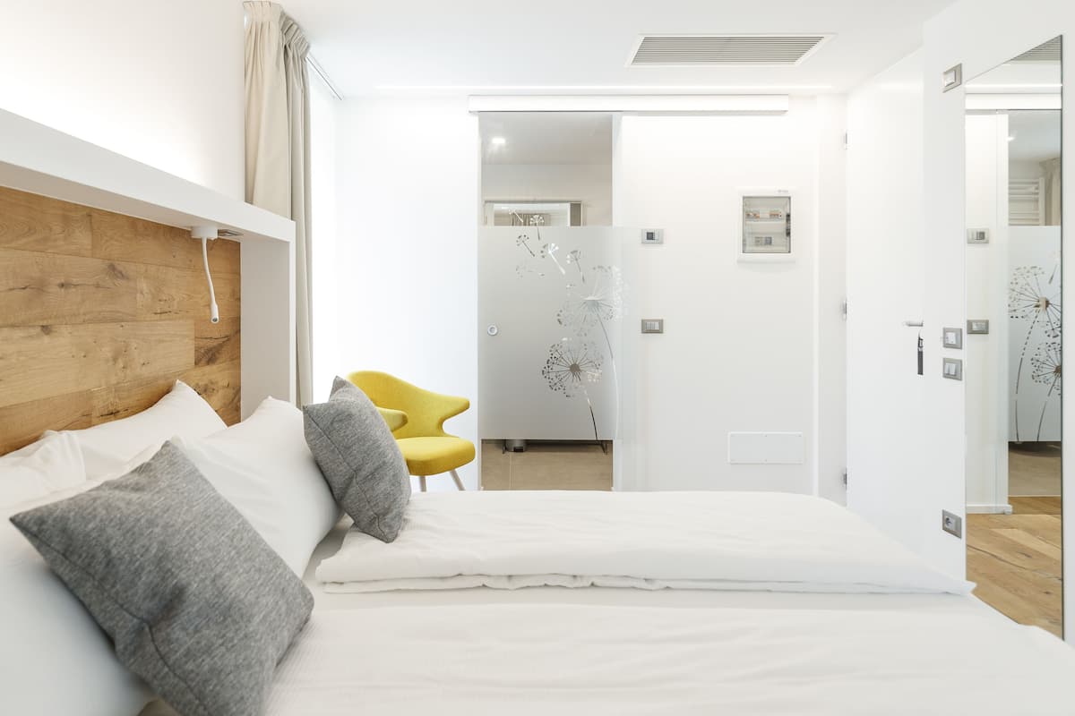 A modern bedroom with a wooden headboard, a bed with white bedding, a yellow chair, and a white door with a dandelion wall decal.