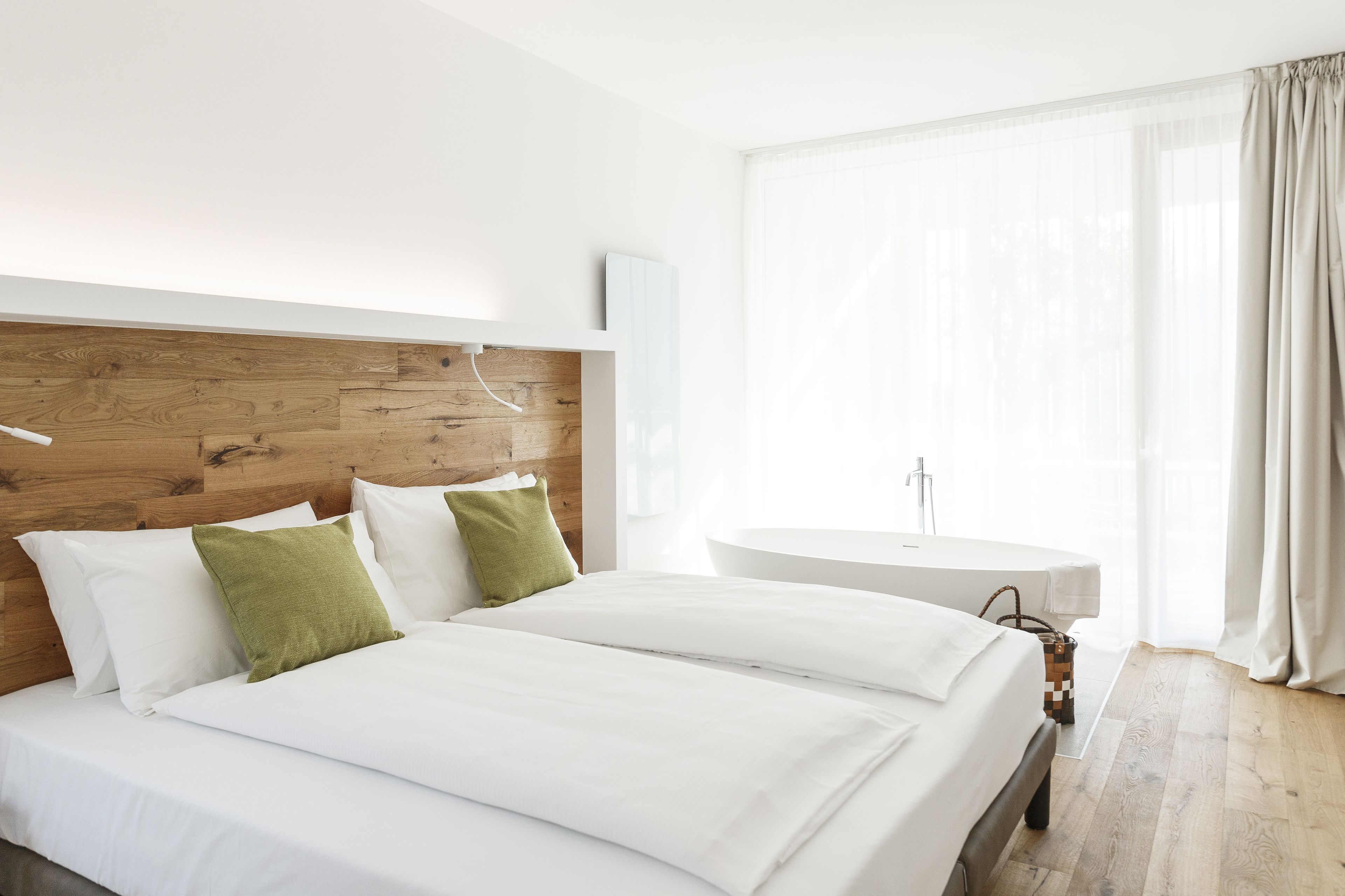 Una camera d'albergo moderna con una testiera in legno, un letto grande con lenzuola bianche, un comodino rotondo e un'ampia finestra con tende leggere.