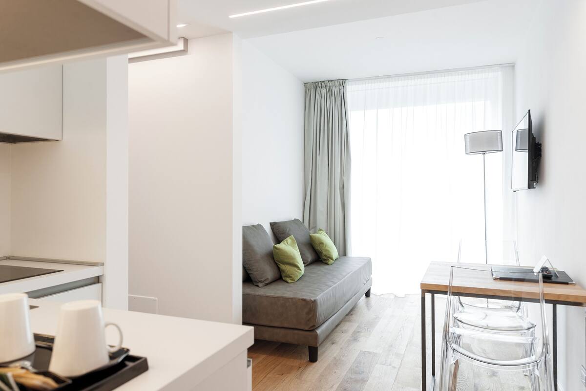 A modern living room with a sofa, a dining table with chairs, a TV mounted on the wall, and a kitchen area with a countertop and appliances.
