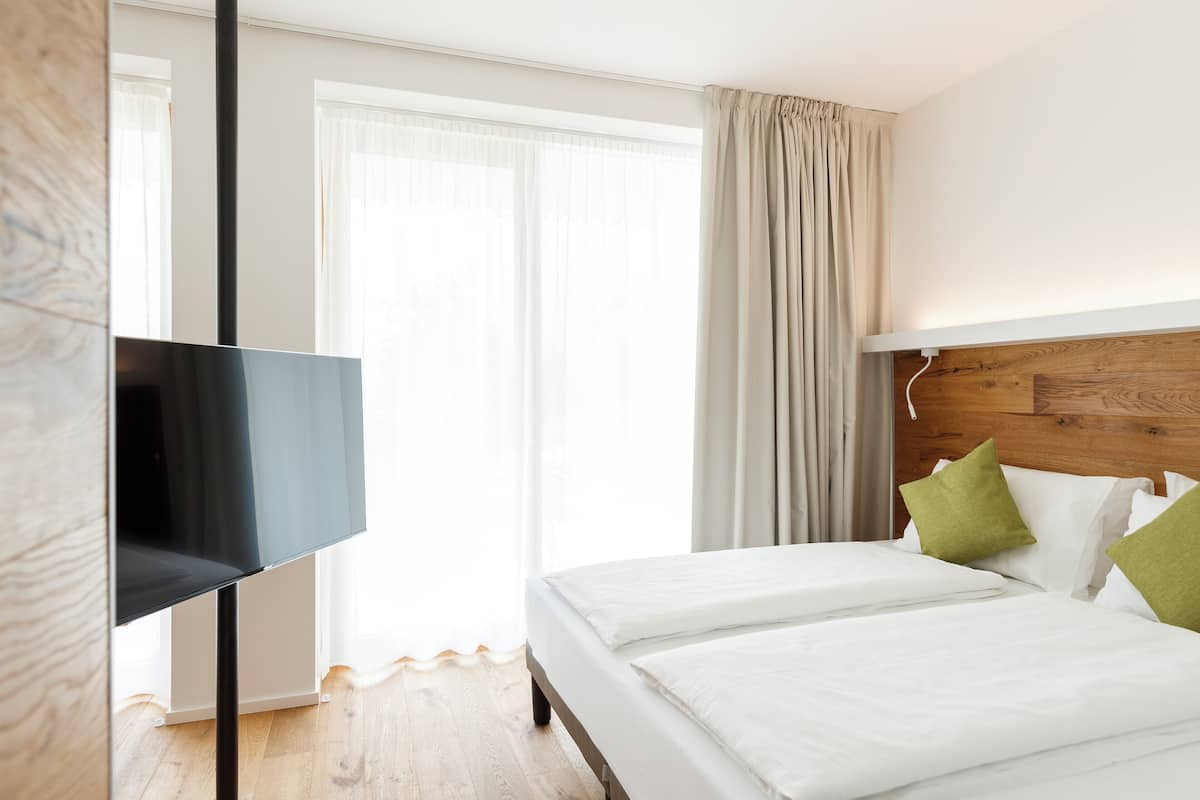 A hotel room with a large flat-screen TV, a wooden headboard, a bed with white bedding and green pillows, and a large window with sheer curtains.