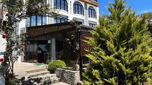 Ein mehrstöckiges Hotel mit überdachter Eingangspartei, Steinstufen und einem großen immergrünen Baum im Vordergrund.