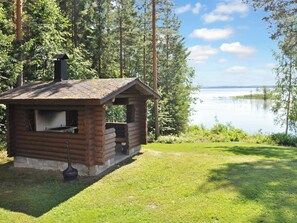 A small wooden cabin with a chimney, located near a lake surrounded by trees.