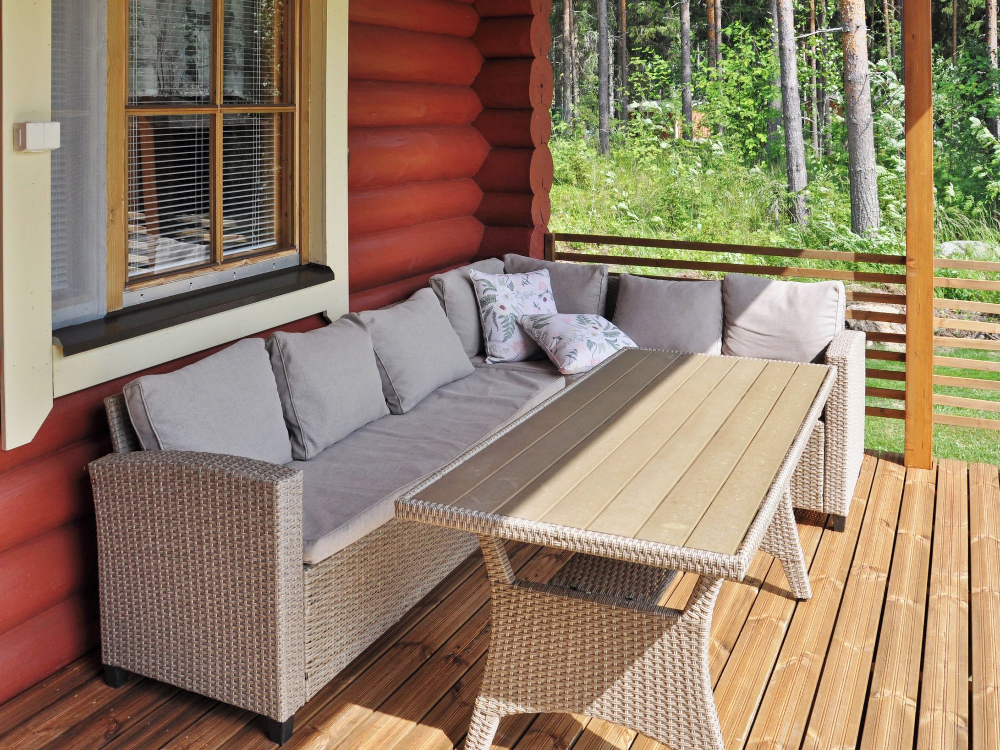 Une terrasse en bois avec un canapé en osier et une table, une fenêtre avec des stores et une vue sur une forêt.