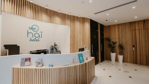 A hotel reception area with a wooden reception desk, a logo on the wall, and potted plants. - Hai Hotel (Larnaca)