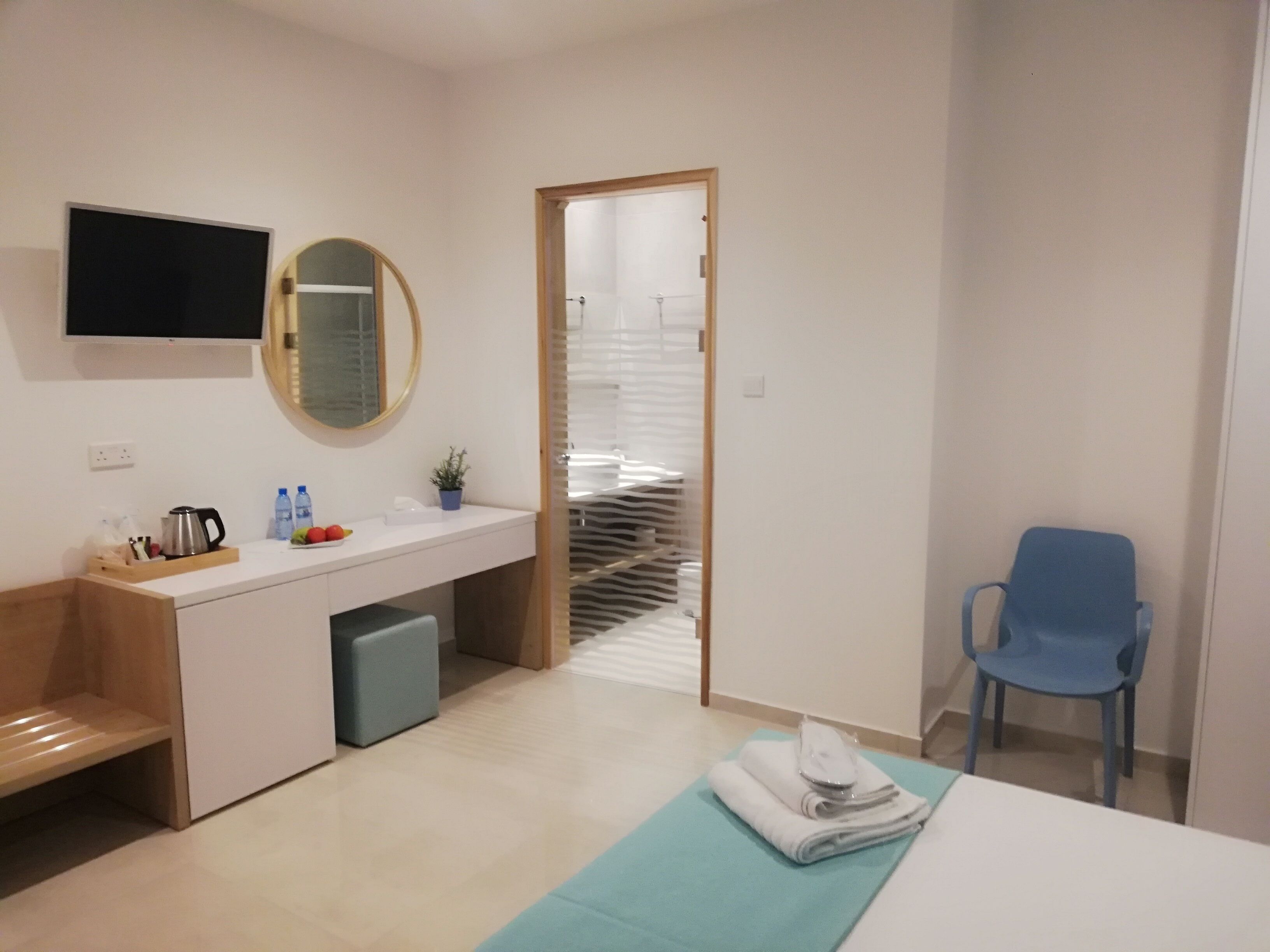 A modern hotel room with a wooden desk, a flat-screen TV, a mirror, a sink, a blue chair, and a bed with a blue bedspread.
