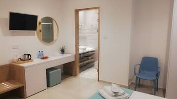 A modern hotel room with a wooden desk, a flat-screen TV, a mirror, a sink, a blue chair, and a bed with a blue bedspread.