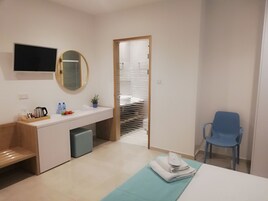 A modern hotel room with a wooden desk, a flat-screen TV, a mirror, a sink, a blue chair, and a bed with a blue bedspread.