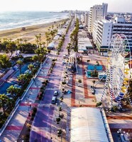 Vista aérea de una zona costera urbana con playa, palmeras, noria y una gran cancha deportiva cubierta.