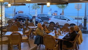 An outdoor restaurant with wicker furniture and hanging lanterns, overlooking a beach with parked cars and people walking. - Hai Hotel (Larnaca)