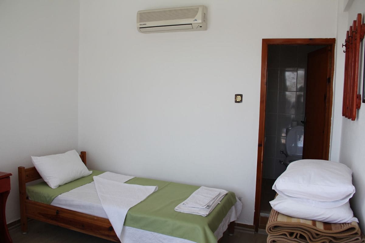 Une petite chambre avec un lit simple, une couvre-lit verte, des oreillers blancs et une serviette blanche sur le lit. On trouve une table de chevet en bois sur la gauche, un climatiseur fixé au mur au-dessus du lit, et une porte donnant accès à la salle de bain.