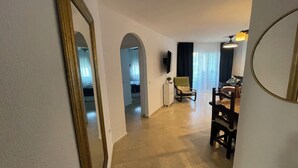 A hallway with a mirrored wall, a seating area with a chair and a table, and a room with a bed and curtains. - Apartment Las Brisas (Mijas-Costa)