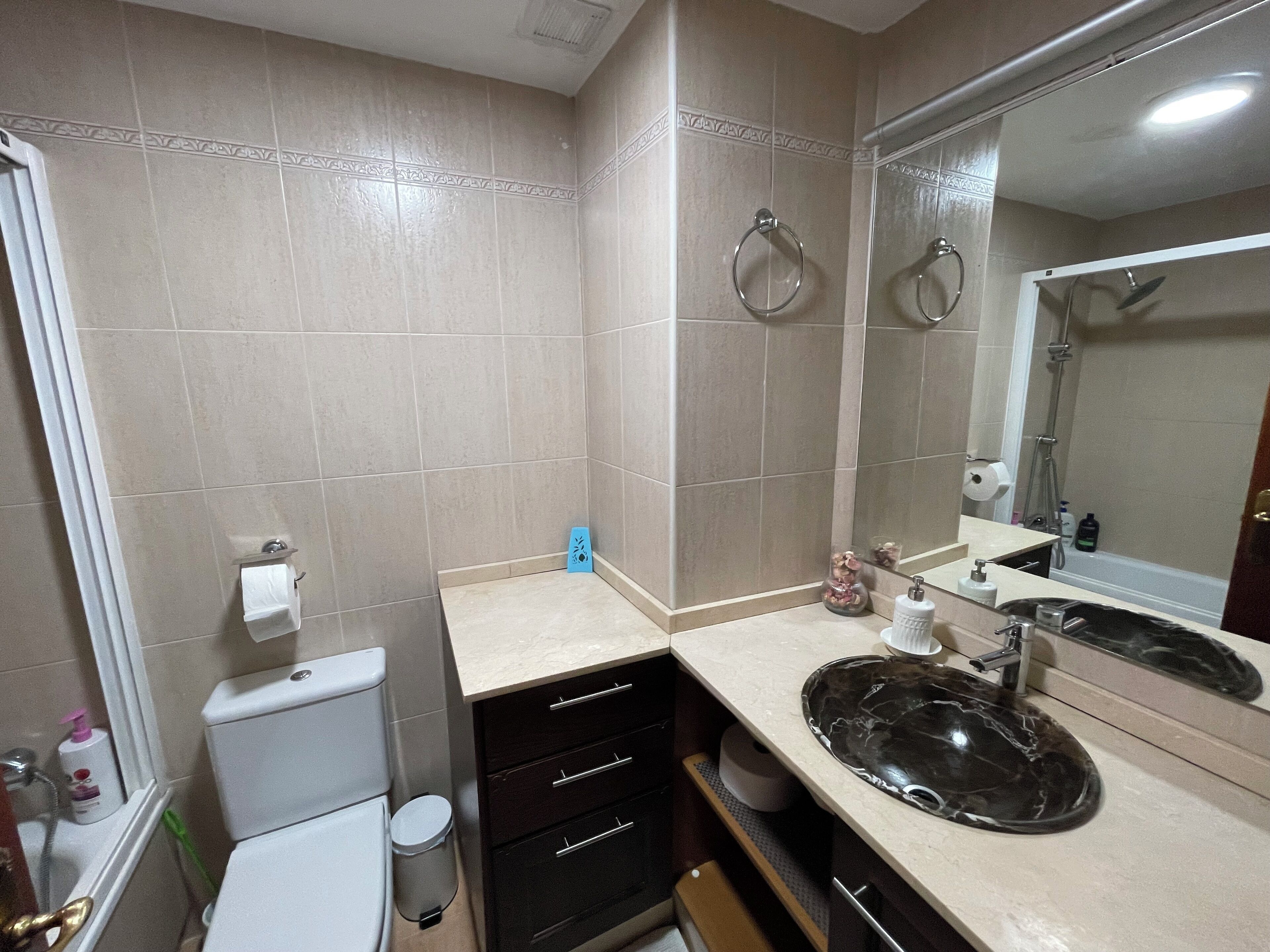 A bathroom with beige tiles, a toilet, a sink with a black marble basin, a mirror, and a shower area.