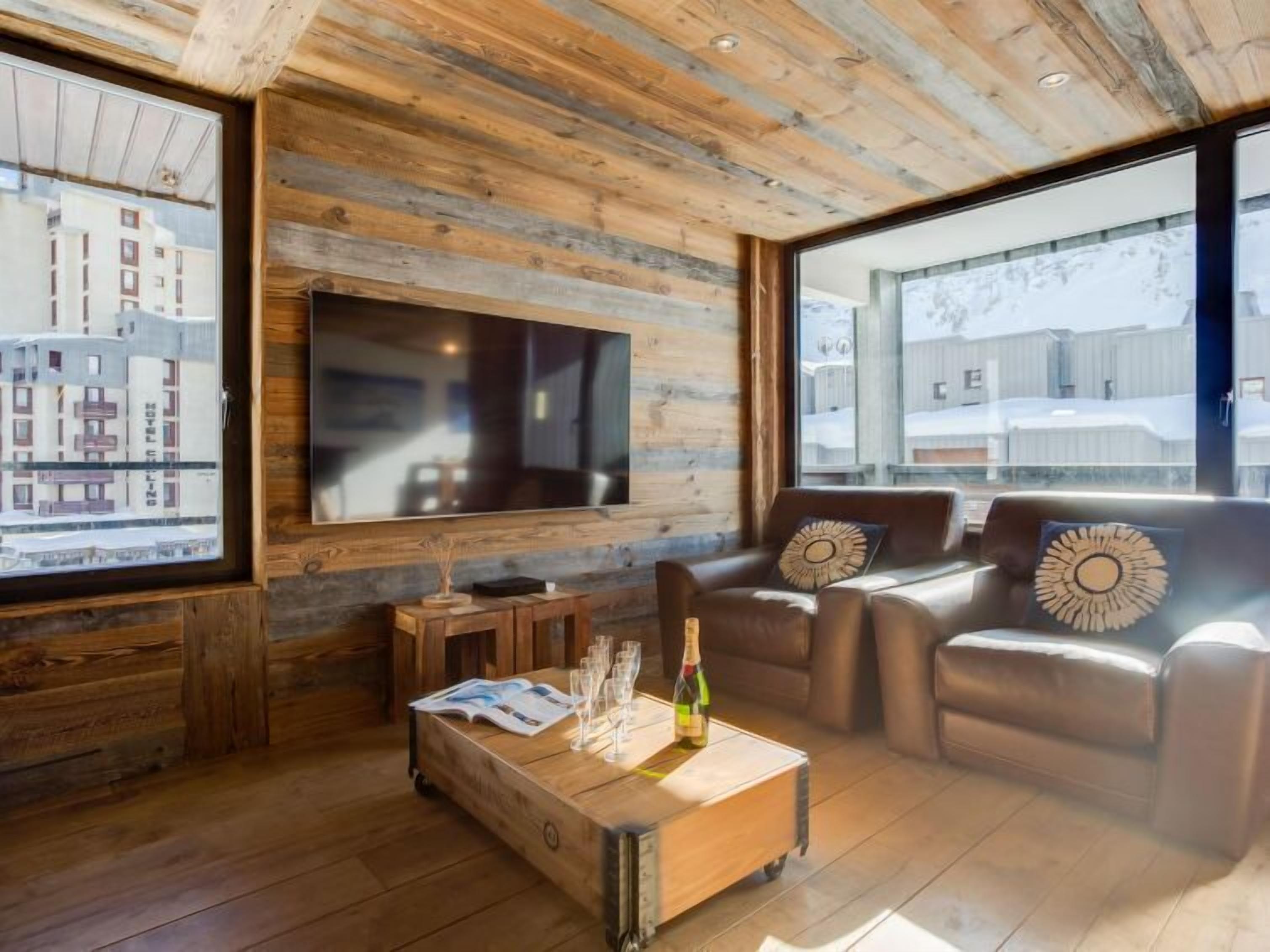 A cozy living room with a large TV, leather sofas, wooden coffee table, and a view of a snowy landscape.