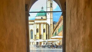Un couloir en arc surmonté d’une voûte, dont le sol est pavé de pierres, mène à une place où se trouvent un bâtiment historique et une tour de l’horloge.
