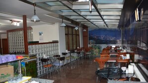 A restaurant with a high ceiling, glass panels, and a mural of a cityscape. Tables and chairs are arranged for dining. - Sadikoglu Oteli (Artvin)