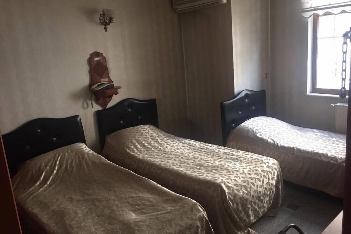 A room with two single beds, a small table, a wall-mounted heater, and a window with a curtain.