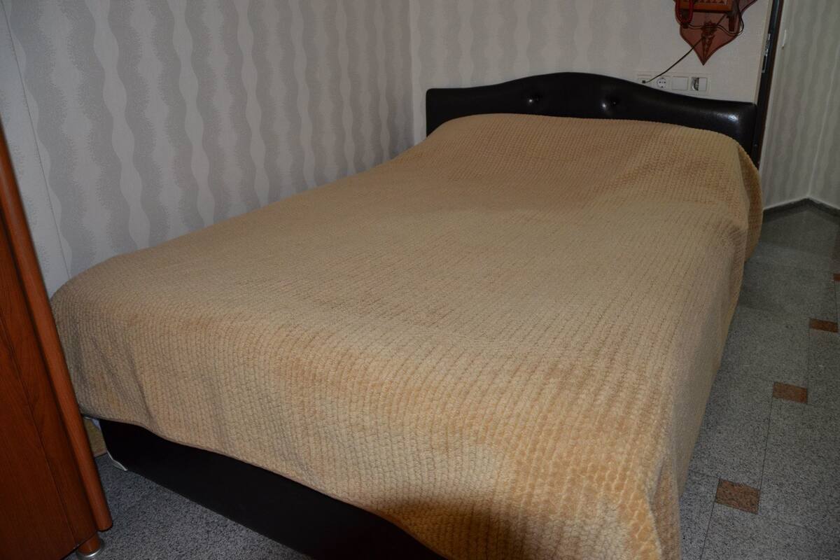 A single bed with a beige blanket in a room with patterned wallpaper and a tiled floor.
