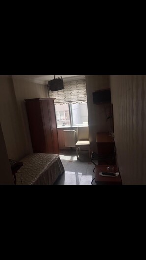 A small, dimly lit room with a bed, a wooden wardrobe, a radiator, a chair, and a television on a stand. - Sadikoglu Oteli (Artvin)