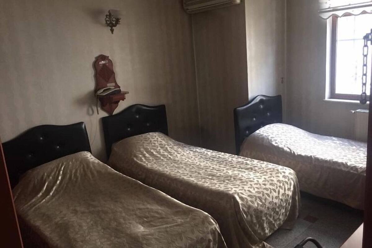 A room with two single beds, a wall-mounted heater, a window with a curtain, and a wall-mounted light fixture.