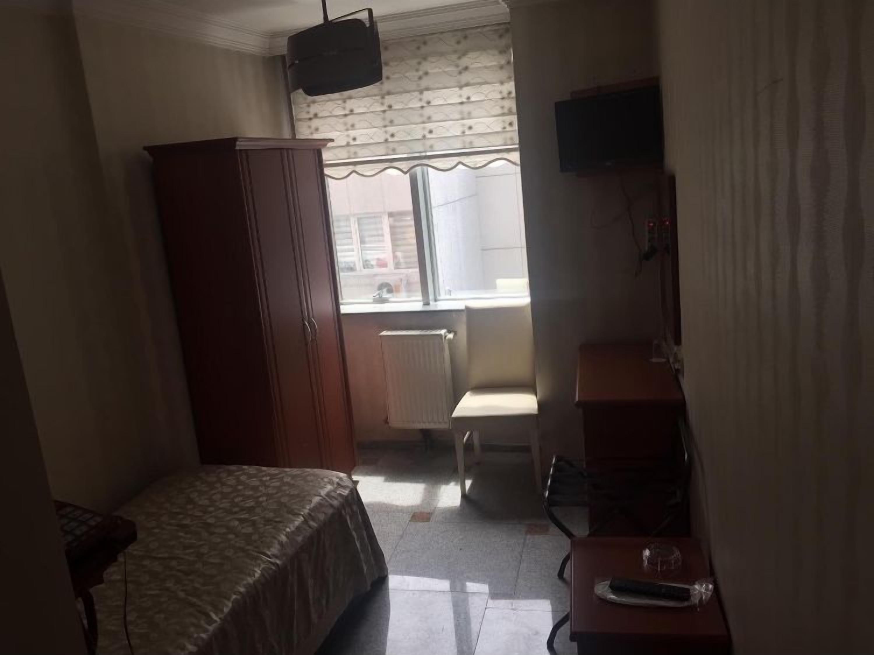 A small, dimly lit room with a bed, a wooden wardrobe, a radiator, a chair, and a television on a stand.