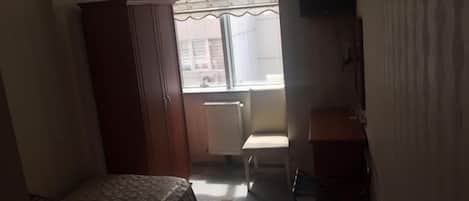 Una habitación pequeña y tenuemente iluminada, con una cama, un armario de madera, un radiador, una silla y un televisor sobre un pedestal.
