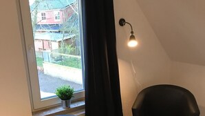 A room with a black leather chair, a small white stool, and a window with a view of houses and greenery. - Kleine Möwe (Dornum)