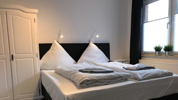 A neatly made bed with pillows and a blanket, a white wardrobe, and a window with plants. - Kleine Möwe (Dornum)