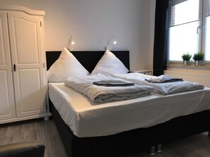 A neatly made bed with pillows and a blanket, a white wardrobe, and a window with plants. - Kleine Möwe (Dornum)