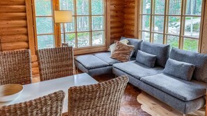 L’intérieur d’un chalet chaleureux, avec une grande fenêtre, un canapé gris, des fauteuils en osier et une table blanche.