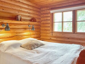 A wooden cabin room with a bed, two wall-mounted lamps, and a window with a view of trees.
