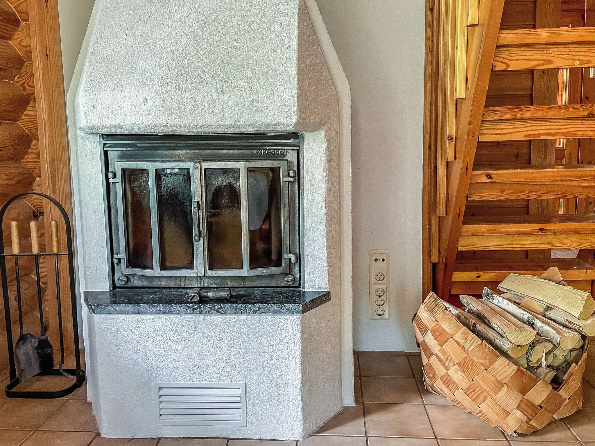 A corner of a room with a white textured wall, a wooden staircase, and a basket of firewood.