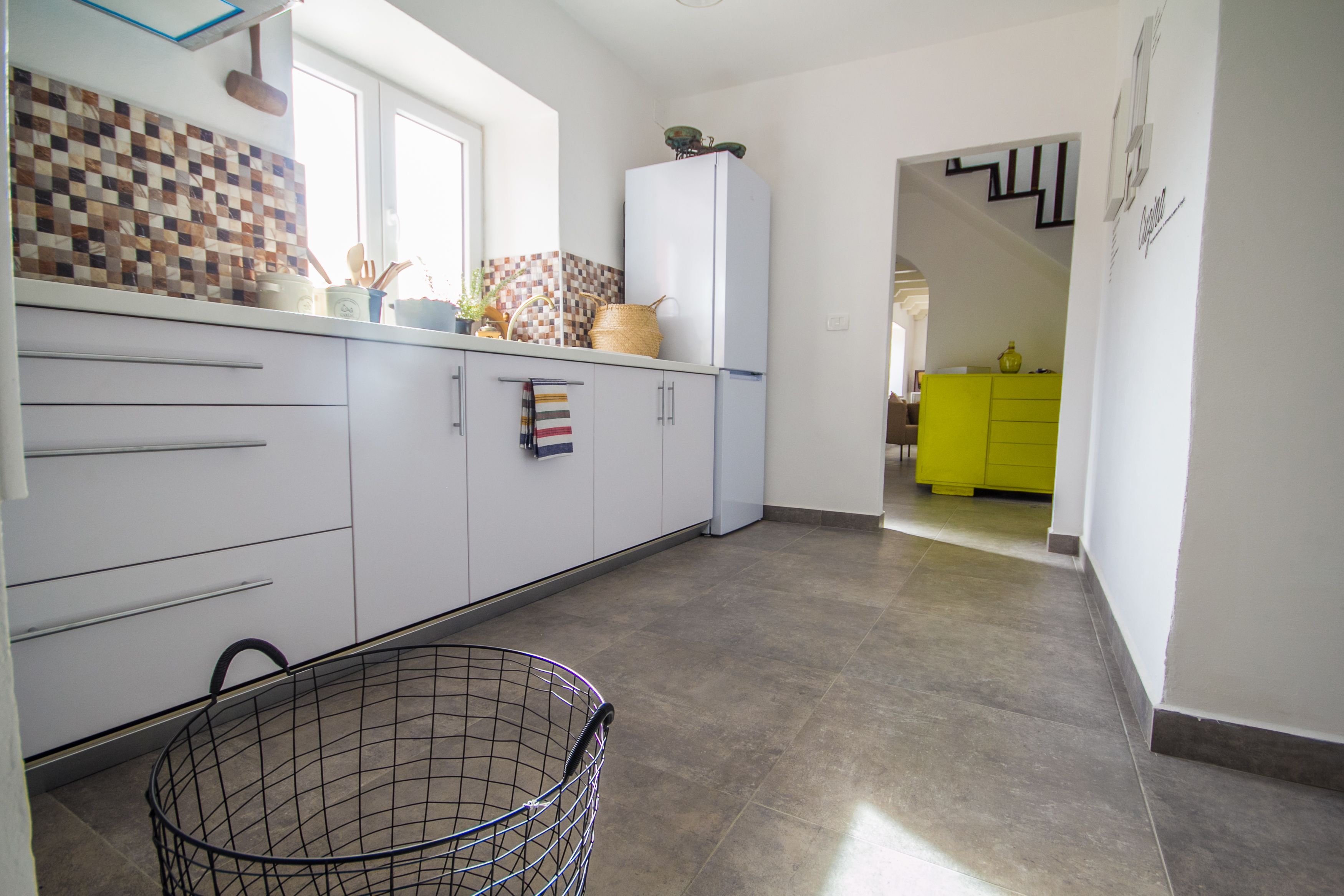 A kitchen with white cabinets, a tiled backsplash, and a window. There is a basket in the foreground and a bright yellow cabinet in the background.