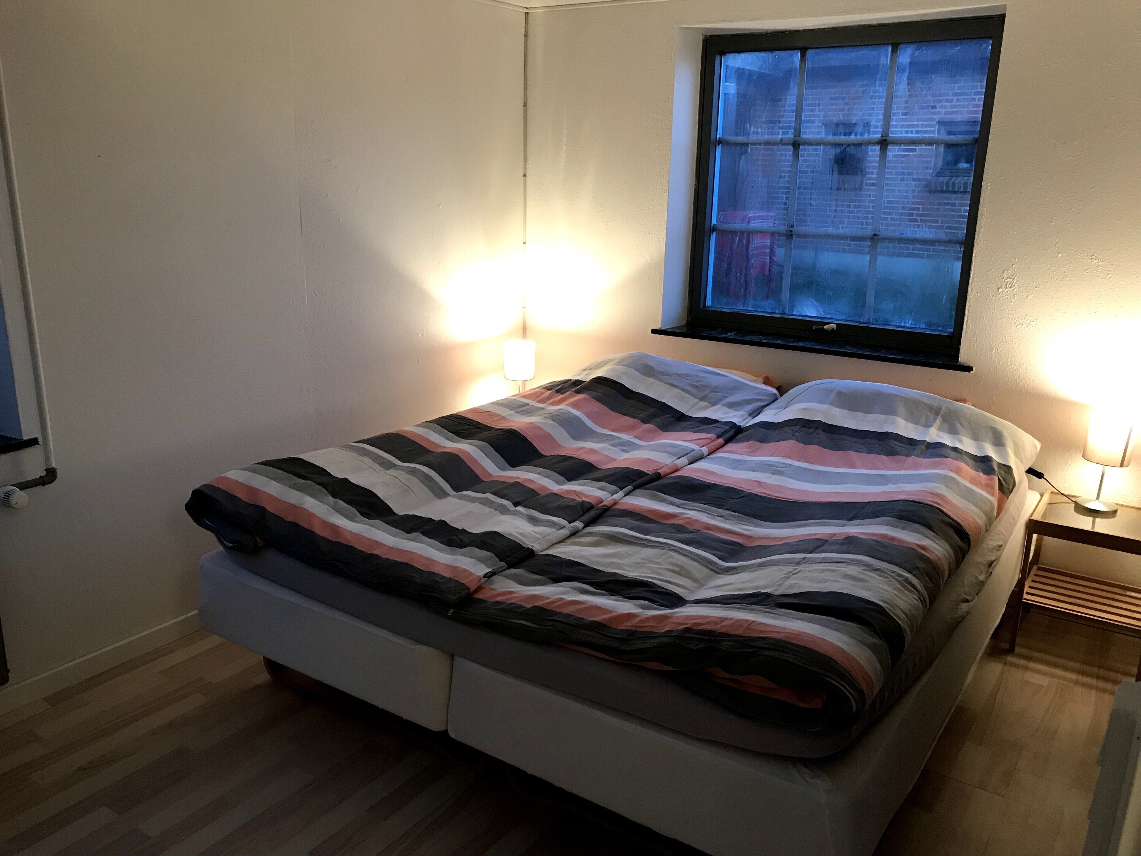 Een eenpersoonskamer met een gestreept beddegoed, twee bedlampen, een raam met uitzicht op gebouwen en een houten vloer.