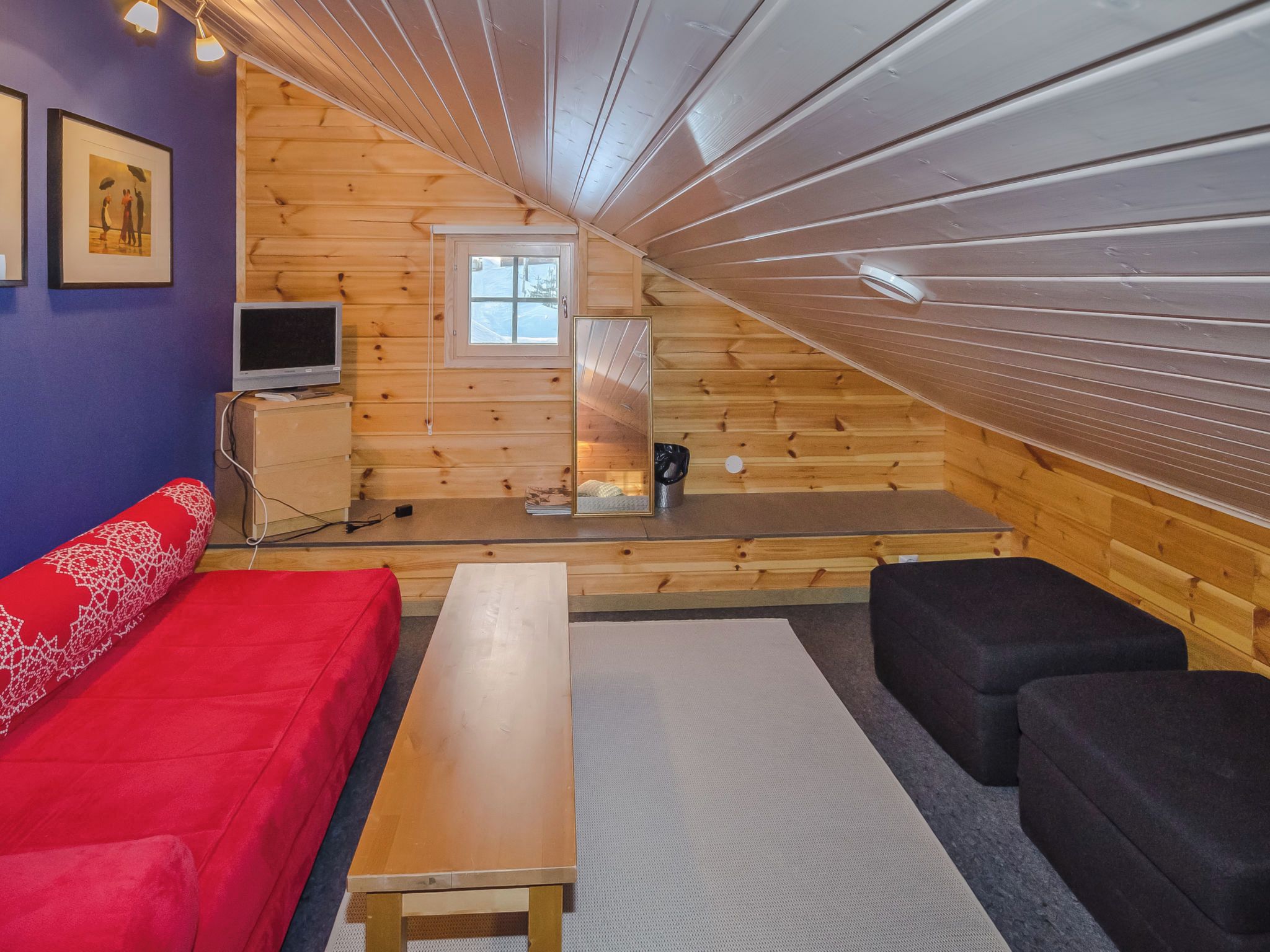 Ein gemütliches Dachzimmer mit einer hölzernen Schräge, einem roten Sofa, einem schwarzen Sessel, einem Holztisch und einem Fernseher auf einer Kommode.